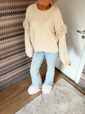 Beige stickad tröja med fransdetaljer - Säljer en beige stickad tröja med oversized passform och fransdetaljer längs ärmarna. Tröjan har rund halsringning och långa, vida ärmar. Perfekt för dig som gillar en avslappnad och trendig stil.