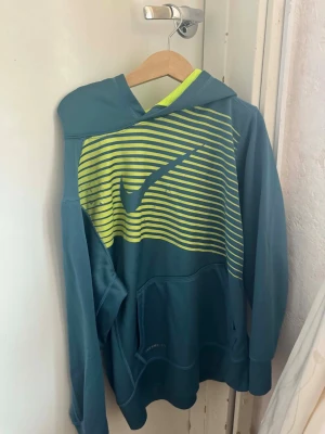 Blå hoodie från Nike med limegula ränder - Snygg blå hoodie från Nike med limegula horisontella ränder och stor Nike-logga på bröstet. Hoodien har huva, magficka och ribbade muddar. Perfekt för dig som gillar sportig stil och vill sticka ut lite extra. Materialet känns mjukt och bekvämt.