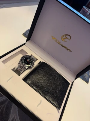 Silverfärgad klocka från Charme - Stilren klocka från Chitime med silverfärgad länk i metall och svart urtavla. Klockan har tydliga markeringar, röd sekundvisare och klassisk design. Perfekt accessoar för dig som vill ha en enkel men snygg look. Plånbok ingår även 