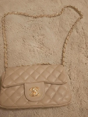 Beige quiltad axelväska - Supersnygg beige axelväska med klassiskt quiltat mönster och guldfärgad kedja. Väskan har det ikoniska CC-spännet framtill och är gjord i glansigt läder. Perfekt för dig som vill ha en tidlös och lyxig accessoar.