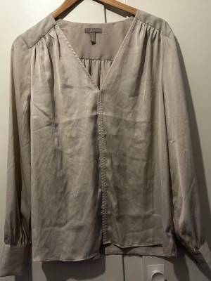 Beige v-ringad blus från H&M - Stilren beige blus från H&M i mjukt, glansigt material. Blusen har v-ringning, långa ärmar med breda manschetter och lätt rynkade axlar för en avslappnad men chic look. Perfekt att matcha med jeans eller kjol.