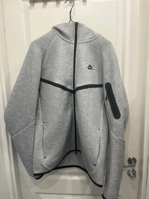 Grå Nike Tech Fleece hoodie - Snygg grå Nike Tech Fleece hoodie med svart dragkedja och detaljer. Har en huva, fickor framtill och en extra ficka med dragkedja på ärmen. Tillverkad i mjukt och stretchigt material som ger en modern och sportig look. Perfekt för dig som gillar streetwear.