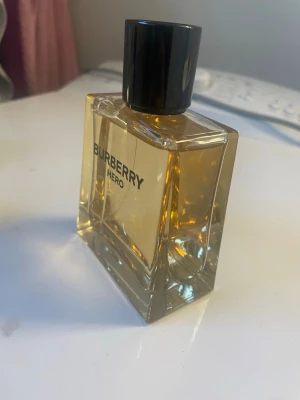 Burberry Hero Eau de Parfum - Burberry Hero är en elegant parfym i en stilren, fyrkantig glasflaska med svart lock. Doften har en varm, gyllene färg och flaskan har tydlig Burberry-logga på framsidan. Perfekt för dig som vill ha en klassisk och modern doft i badrumshyllan.