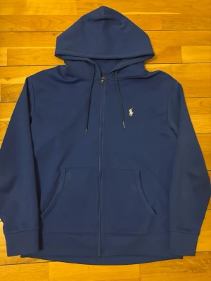 Blå hoodie från Polo Ralph Lauren - Snygg blå hoodie med dragkedja från Polo Ralph Lauren. Klassisk design med huva och dragsnören, två fickor framtill och den ikoniska vita logon broderad på bröstet. Perfekt för en avslappnad stil och enkel att matcha med jeans eller joggers.