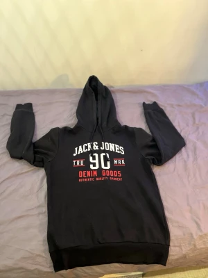 Svart hoodie från Jack & Jones - Svart hoodie från Jack & Jones med tryck i vitt och rött på bröstet där det står 'Jack & Jones 90 Denim Goods'. Klassisk huva med dragsko och ribbade muddar vid ärmslut och nederkant. Perfekt för en avslappnad och cool stil.