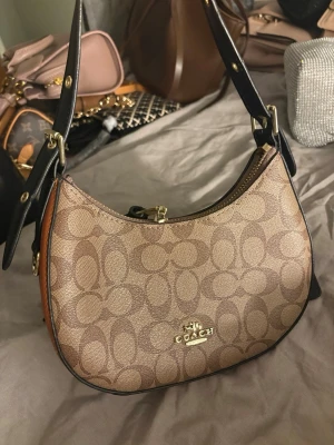 Coach beige - Snygg handväska från Coach i beige med klassiskt monogrammönster och guldfärgad logga framtill. Väskan har svarta justerbara axelremmar och bruna detaljer på sidorna. Tillverkad i tåligt canvasmaterial med dragkedja upptill.   Dragkedjan är lite sönder 