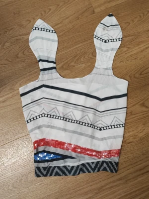 Mönstrad crop top med paljetter - Unik crop top med breda axelband som knyts runt nacken och spetsiga detaljer. Toppen är vit med svarta, grå och blå grafiska mönster samt röda och blå paljetter. Dragkedja bak för enkel påklädning. Perfekt för dig som vill sticka ut med en kreativ och färgglad stil.