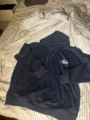 Mörkblå hoodie från Stüssy - Säljer en mörkblå hoodie från Stüssy med klassisk logga i vitt på bröstet. Hoodien har huva med dragsko och en stor magficka. Tillverkad i mjukt bomullsmaterial, perfekt för en avslappnad streetwear-look.