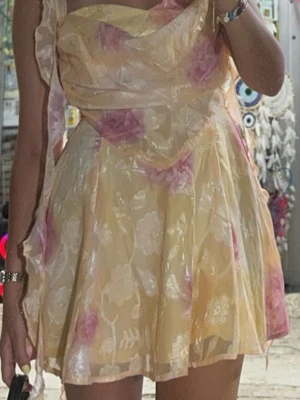 Blommig kortklänning med volang - Söt kortklänning i ljusgult med rosa och vita blommor. Klänningen har volangdetaljer, omlottliknande överdel och öppen rygg med tunna axelband. Tillverkad i ett lätt, transparent material som ger en romantisk känsla. Har dragkedja baktill också 