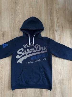 Mörkblå Superdry hoodie med tryck - Mörkblå hoodie från Superdry med stort vitt och blått tryck på bröstet och en blå siffra på ärmen. Klassisk känguruficka framtill och vita dragsnören i huvan. Perfekt för en avslappnad och cool stil. Storleken är S men sitter som M