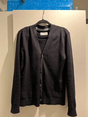 Svart kofta från Pier One - Stilren svart kofta från Pier One med knäppning framtill och ribbade muddar vid ärmslut och nederkant. Klassisk V-ringning och mjukt material som passar perfekt till lager-på-lager-stil. Skriv gärna om ni har frågor 