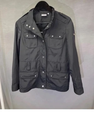 Svart Fieldjacka - Stilren svart feildjacka/vindjacka med fyra framfickor och tryckknappar. Jackan har hög krage, axelklaffar och justerbara ärmslut. Materialet är lätt och vindtätt, perfekt för blåsiga dagar. Passar dig som gillar en enkel men cool look. Det är en jacka från Esselle i storlek S skriv om ni har några funderingar. Priset kan diskuteras 