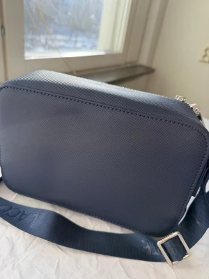 Mörkblå axelväska från Lacoste - Snygg mörkblå axelväska från Lacoste i tåligt textilmaterial med struktur. Väskan har justerbar axelrem med Lacoste-logga och silverfärgade dragkedjor med krokodilemblem. Perfekt storlek för det viktigaste och stilren design.