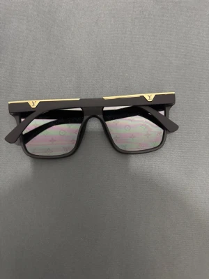 Louis Vuitton svarta solglasögon - Snygga svarta solglasögon från Louis Vuitton med spegelglas och ikoniskt LV-monogram över hela linsen. Rektangulär form med gulddetaljer på ovansidan av bågen för en exklusiv look. Perfekta för dig som vill sticka ut och addera lyx till din stil.