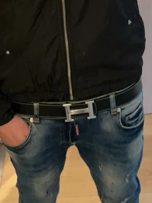 Svart skärp med H-spänne - Snyggt svart bälte med ett stort H-format metallspänne och vita kontrastsömmar. Perfekt accessoar för att lyfta din outfit med en stilren och klassisk look. Bältet har en slät yta och är tillverkat i skinn.