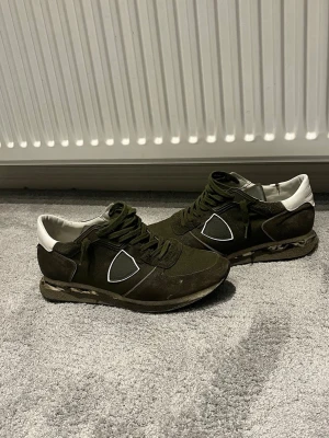 Olivgröna sneakers från Philippe Model - Snygga olivgröna sneakers från Philippe Model med vita detaljer och logga på sidan. Ovandelen är i mocka och textil, med vita partier vid hälen och en unik sula med mönster. Perfekta för dig som gillar streetwear och vill sticka ut.har en lite. Skada i inre sidan som man ser på bilden märks ej av och har varit där jätt länge så är nog inge risk att den går i sönder mer 