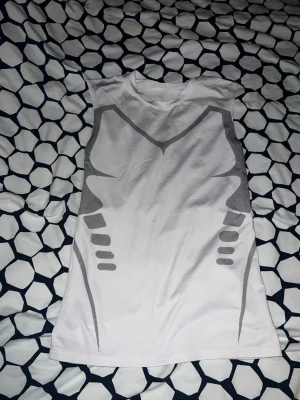 Vit ärmlös linnetopp med grått mönster - Säljer en vit ärmlös linnetopp med grått grafiskt mönster både fram och bak. Toppen har en sportig look och är tillverkad i ett lätt och stretchigt syntetmaterial som passar perfekt till träning eller en aktiv vardag.