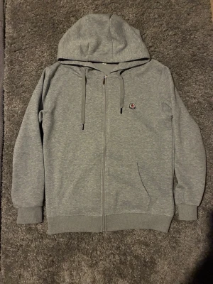 Moncler zip hoodie grå - Snygg grå mocler hoodie med dragkedja framtill, huva med dragsko och ribbade muddar. Liten Moncler-logga på bröstet ger en clean look. Perfekt för dig som gillar stilrena och bekväma plagg. Tillverkad i mjukt bomullsmaterial.