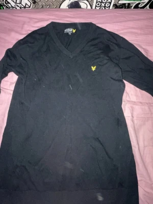 Svart stickad tröja från Lyle & Scott - Svart stickad tröja från Lyle & Scott med V-ringning och klassisk gul logga broderad på bröstet. Tröjan har långa ärmar och en stilren, enkel design som passar perfekt till jeans eller chinos.