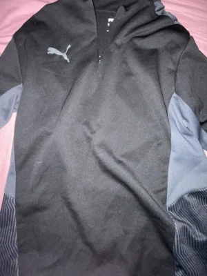 Svart hoodie från Puma med huva - Säljer en svart hoodie från Puma med gråa detaljer på ärmarna och en snygg Puma-logga på bröstet. Hoodien har dragkedja upptill och är tillverkad i ett mjukt, sportigt material som passar perfekt till chill eller träning.