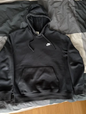 Svart hoodie  Nike med logga - Säljer en svart hoodie från Nike med vit broderad logga på bröstet. Hoodien har huva med dragsko och en stor magficka framtill. Tillverkad i mjukt material och har ribbade muddar vid ärmar och nederkant. Perfekt för en avslappnad streetwear-look.