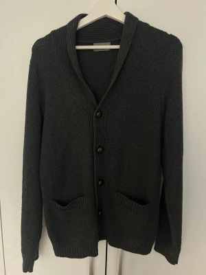 Mörkgrå stickad kofta med knappar från Grand Frank - Mysig mörkgrå stickad kofta med bruna knappar framtill och två fickor. Koftan har ribbstickade muddar och en bred krage som ger en avslappnad look. Perfekt att slänga på sig när det är kyligt ute.