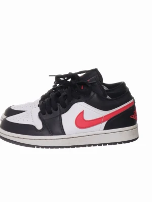 Nike Air Jordan 1 Low svart/vit/röd - Snygga Nike Air Jordan 1 Low sneakers i svart, vitt och rött. Skorna har klassisk låg siluett, svart läder på ovandelen, vit tåbox och sidor samt röda detaljer som swoosh och logga på hälen. Perfekta för dig som gillar streetwear och ikonisk design.