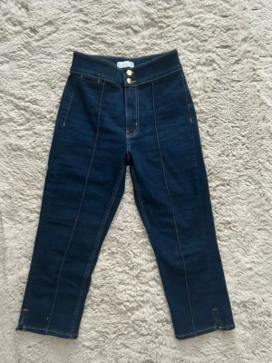 Mörkblå capri jeans - Snygga mörkblå jeans med hög midja och raka ben. Byxorna har dekorativa sömmar framtill, två guldknappar i midjan och fejkade bakfickor. Klassisk denimlook med modern twist, perfekta till sneakers eller boots. 