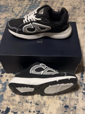 Svarta Dior sneakers med vita detaljer - Snygga svarta Dior sneakers med vita och grå detaljer samt tydlig Dior-logga på sidan och sulan. Skorna har en chunky vit sula och vita skosnören. Ovandelen är i mesh och mockaliknande material för en sportig och modern look.