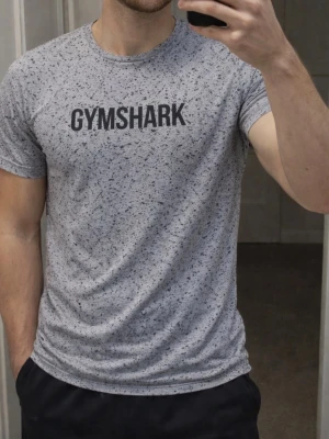Gymshark t-shirt - Snygg grå t-shirt från Gymshark med svart logga på bröstet. T-shirten har korta ärmar och ett unikt prickigt mönster över hela tyget. Perfekt för dig som gillar sportig och trendig stil. Storlek: S