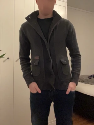 Mörkgrå kofta med knappar och fickor - Säljer en mörkgrå kofta från river island med bred krage, knäppning framtill och två stora fickor med lock. Koftan har ribbade muddar och är tillverkad i ett mjukt stickat material. Perfekt för lager-på-lager och snygg till jeans.