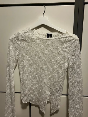 Vit spetsblus från Vero Moda - Säljer en vit långärmad blus från H&M i genombruten spets med blommigt mönster. Toppen har rund halsringning och är figurnära med en lätt transparent look. Perfekt att styla lager på lager eller med ett linne under.