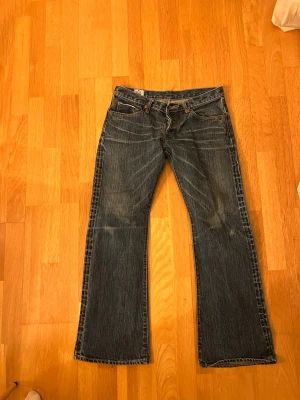 Blå low waist bootcut jeans - Säljer ett par klassiska blå bootcut jeans  med snygga slitningar och kontrastsömmar. Jeansen har fem fickor, låg midja och är tillverkade i kraftig denim. Perfekta för en avslappnad och trendig look. Innerbenslängd: 74 cm. Midjemått rakt över: ca 40 cm