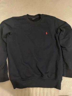 Svart sweatshirt från Ralph Lauren - Klassisk svart sweatshirt från Ralph Lauren med rund halsringning och den ikoniska röda logotypen broderad på bröstet. Tröjan har ribbade muddar vid ärmslut och nederkant, perfekt för en clean och stilren look.