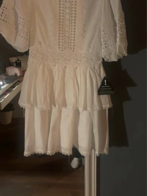 Vit kort klänning med spetsdetaljer - Söt vit kortklänning med volanger och vackra spetsdetaljer på ärmar och överdel. Klänningen har trekvartsärm och omlottliknande spetsmönster framtill. Perfekt för sommarens alla tillfällen och student.