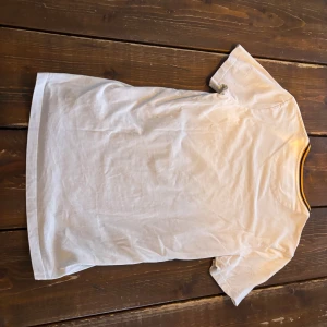 Vit t-shirt från Lyle & Scott - Vit t-shirt från Lyle & Scott med klassisk gul broderad logga på bröstet. T-shirten har rund hals med gul och svart detalj runt kragen och är kortärmad. Perfekt basplagg med stilren design.