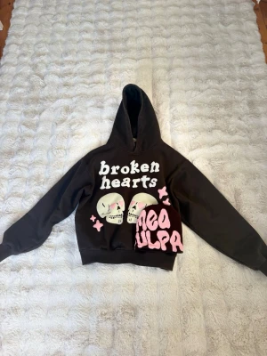 Broken Planet Brun hoodie Broken Hearts  - Mörkbrun hoodie från Broken Planet med stor huva och tryck framtill. Texten 'broken hearts' och två döskallar med rosa detaljer samt rosa stjärnor. Mjuk bomullskänsla och oversize passform. Perfekt för dig som gillar streetwear och statement-plagg.