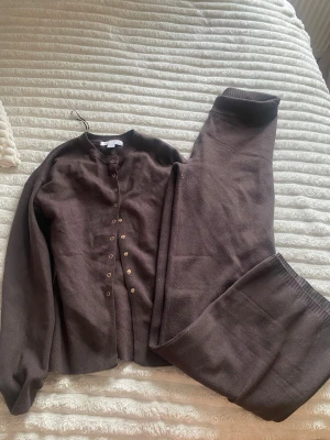 Brunt loungewear-set med knappar - Mysigt brunt loungewear-set med långärmad tröja och matchande byxor. Tröjan har rund halsringning och knäppning framtill med små guldknappar. Byxorna har resår i midjan för extra komfort. Perfekt för chill dagar hemma.