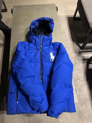 Blå pufferjacka från Polo Ralph Lauren - Snygg blå pufferjacka från Polo Ralph Lauren med stor vit logga på bröstet. Jackan har huva, dragkedja framtill och sidofickor med dragkedja. Insidan är fodrad i mörkblått och har en innerficka. Perfekt för kalla vinterdagar.