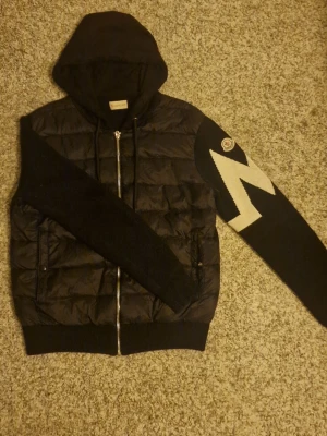 Svart Moncler pufferjacka med huva / moncler tricot cardigan - Säljer en svart pufferjacka från Moncler med huva och dragkedja. Ena ärmen har ett stort vitt blixtmönster och Moncler-logga på axeln. Jackan har quiltad kropp och stickade ärmar för en unik look. Två fickor framtill med knappstängning.