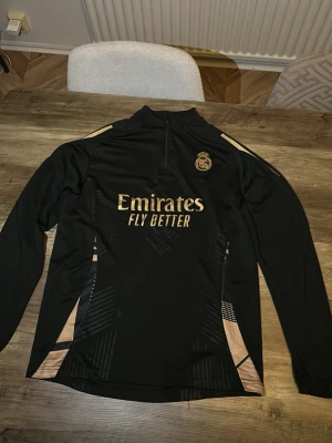 Real Madrid träningströja svart/guld - Snygg långärmad träningströja från Real Madrid i svart med guldiga detaljer och klubbmärke på bröstet. Tröjan har halv dragkedja, trycket 'Emirates Fly Better' och är tillverkad i ett lätt och funktionellt material. Perfekt för fotbollsträning eller sport.