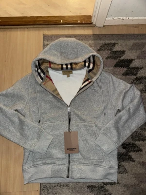 Grå zip hoodie från Burberry - Snygg grå zip hoodie från Burberry med klassiskt rutigt foder i huvan och diskret Burberry-logga på bröstet. Helt ny, säljs för att den är för stor 