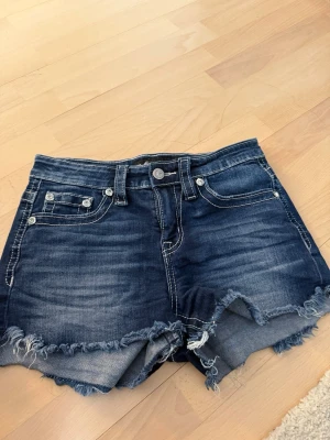 ”Dark wash” jeansshorts med broderi - Säljer ett par mörkblå jeansshorts från Miss Me liknande märke med slitna kanter och snygga broderade detaljer med paljetter på bakfickorna. Klassisk femficksmodell med låg midja och råa benslut. Perfekta för sommaren!
