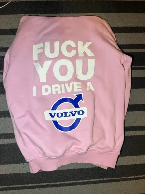 Rosa Volvo hoodie med tryck - Rosa hoodie med stort vitt och blått tryck på ryggen där det står 'FUCK YOU I DRIVE A VOLVO' samt Volvos logga. På framsidan finns också Volvos logga i blått och vitt. Hoodien har klassisk passform och ribbade muddar. Perfekt för dig som vill sticka ut och visa din Volvo-pride.