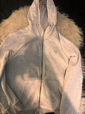 Ljusgrå zip hoodie från Gant - Ljusgrå hoodie från Gant med dragkedja framtill och klassisk huva. Tröjan har fickor på magen och diskret Gant-logga broderad på bröstet. Tillverkad i mjukt sweatshirtmaterial som är perfekt för en avslappnad stil.