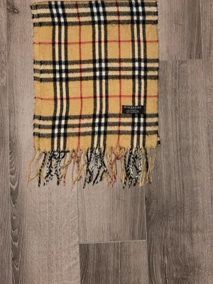 Klassisk beige rutig halsduk Burberry - Säljer en ikonisk beige halsduk från Burberry med klassiskt rutmönster i svart, vitt och rött. Halsduken har fransar i ändarna och är tillverkad av 100% lammull. Perfekt accessoar för att lyfta din outfit.