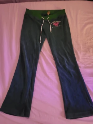 Gröna Juicy Couture mjukisbyxor - Mörkgröna mjukisbyxor från Juicy Couture med bred resår i midjan och vitt dragsnöre. Byxorna har utsvängda ben och en liten logga i rosa och vitt på ena höften. Mjukt velourmaterial som känns riktigt skönt. Byxorna är L men eftersom de är vintage varierar storleken. Längden från midjan till fötterna är ca 95cm och midjan är 39cm bred. Man ser att de är gamla och är litte nötta i midjan men de ser bra ut. Säljer för att storleken inte passar mig. Vill ha sålt snabbt så priset går att diskutera.