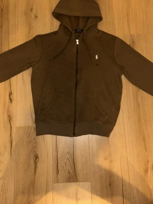 Kahaki/grön hoodie från Polo Ralph Lauren - Snygg khaki/grön hoodie från Polo Ralph Lauren med dragkedja framtill och klassisk vit broderad logga på bröstet. Hoodien har huva med dragsko och två fickor fram. Tillverkad i mjukt bomullsmaterial som känns skönt mot huden. Pris: 600 kr/ pris kan diskuteras vid snabb affär.                 