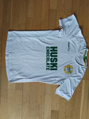 Hammarby matchtröja Craft XS - Vit Hammarby fotbollströja från Craft med gröna detaljer och klubbmärke på bröstet. Tryck med 'Huski Chocolate' på framsidan. Rund hals med grön och vit rand. Perfekt för dig som vill visa ditt stöd på läktaren eller träningen.