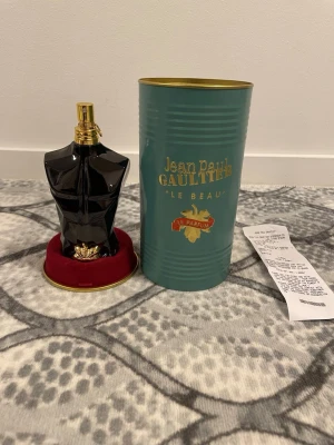Jean Paul Gaultier Le Beau Le Parfum - Snygg parfymflaska formad som en svart manlig torso med gulddetaljer från Jean Paul Gaultier. Kommer i en grön metallburk med guldtext och röd sammetsbotten. Perfekt för dig som gillar unika och lyxiga dofter.